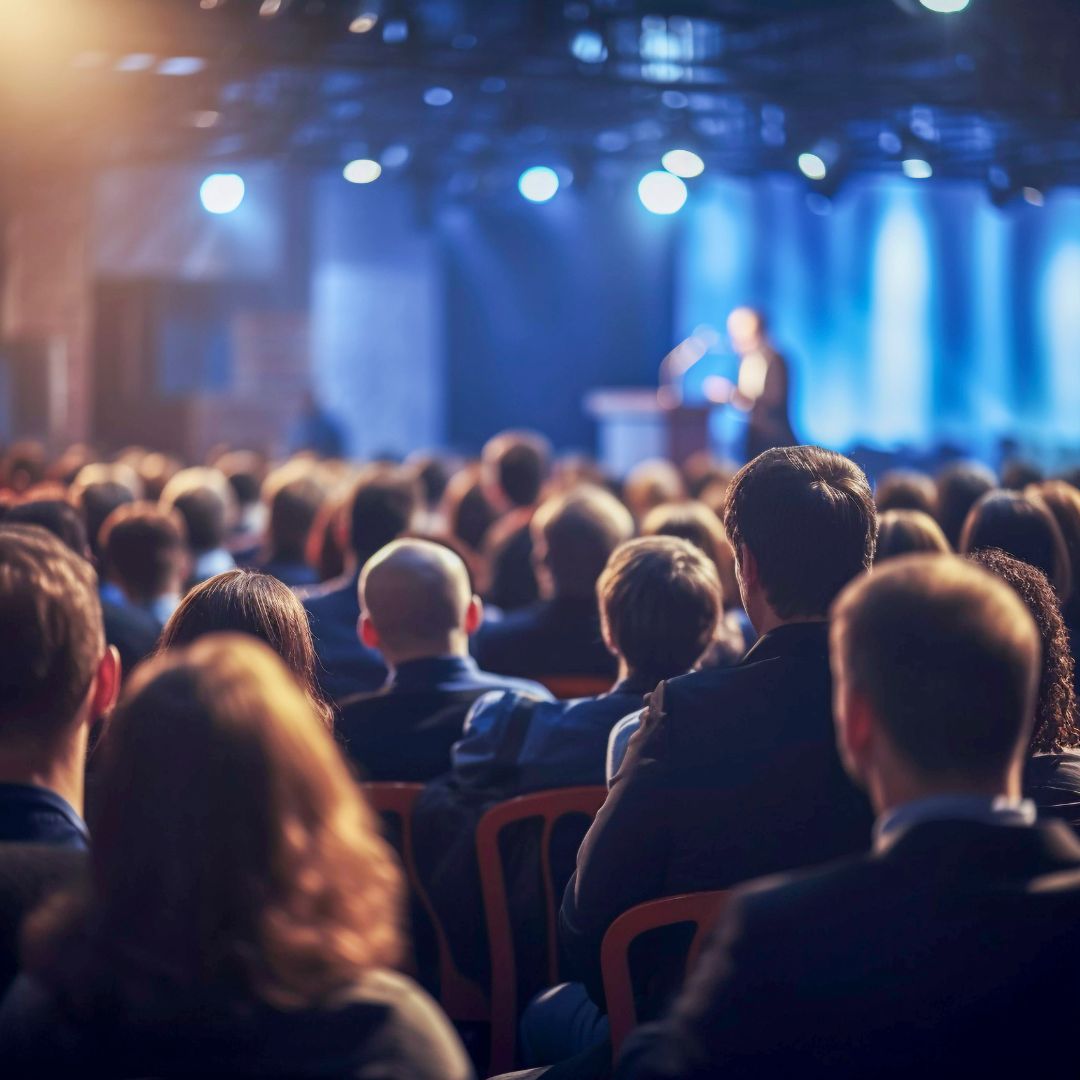 Persone sedute di spalle che ascolto una persona che parla su un palco durante un congresso